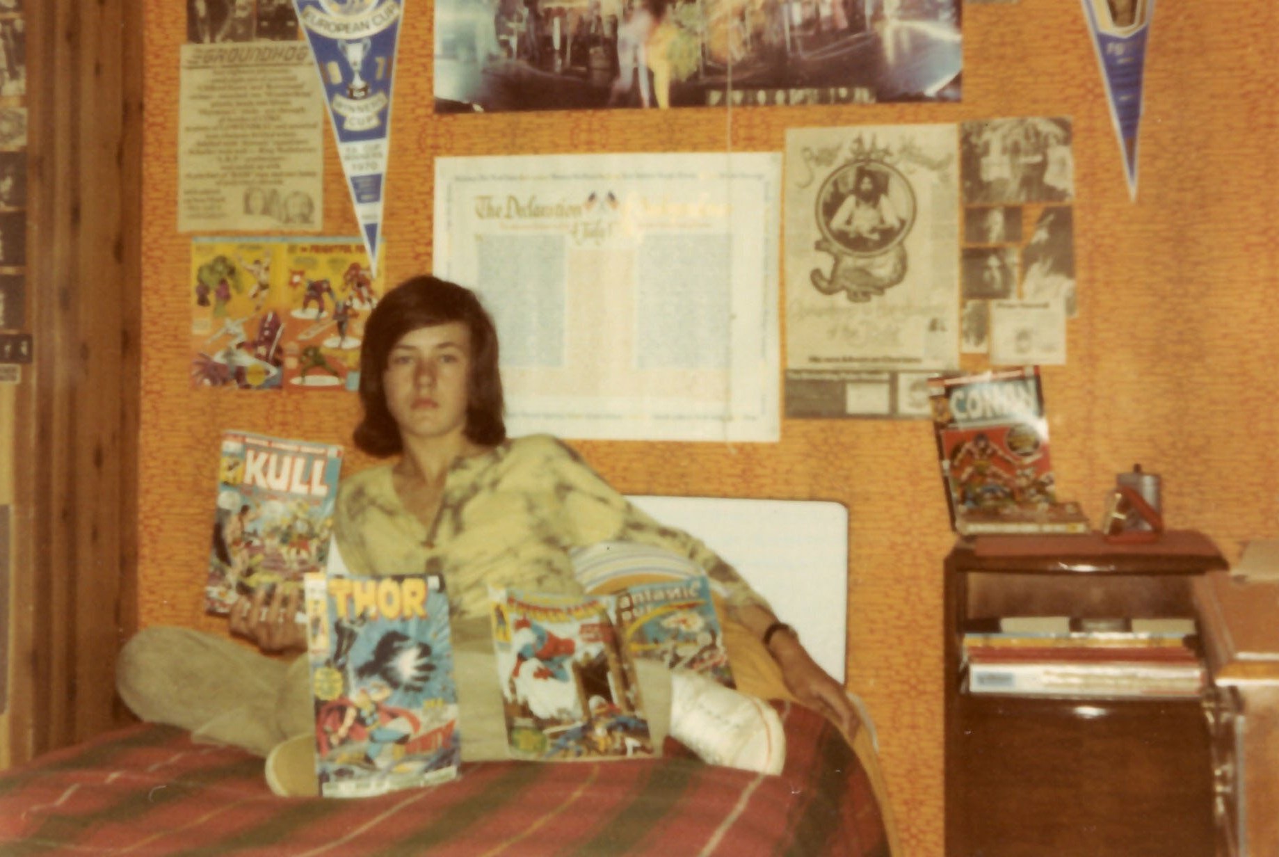 Photo of a teenager sitting on a bed surrounded by comic books and posters.