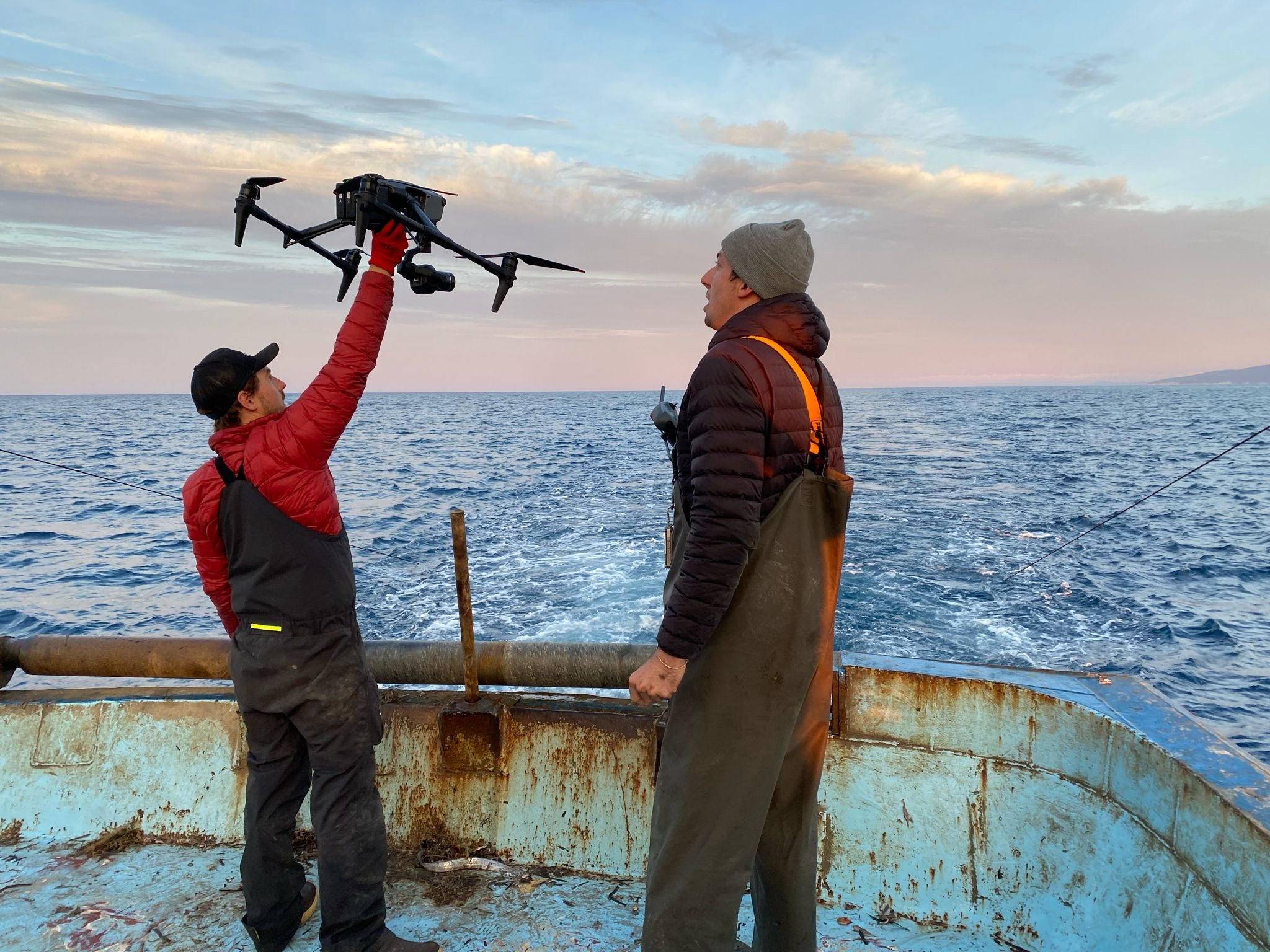 Two individuals on a boat deploy a drone to film an ocean trawler.